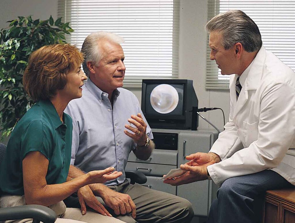 A man in a consultation with a hearing aid specialist, discussing the potential need for a hearing aid. His wife is present, providing supportive assistance during the appointment