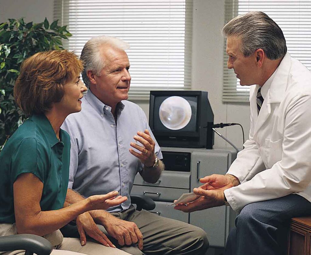 A man in a consultation with a hearing aid specialist, discussing the potential need for a hearing aid. His wife is present, providing supportive assistance during the appointment